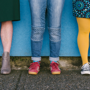 Image of three pairs of legs standing in front of a blue wall. On the left they are wearing a green dress. In the middle someone wearing jeans with red sneakers, finally on the right a person wearing a patterned skirt with bright yellow tights and blue shoes.