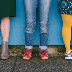 UN/TIED SHOES Image of three pairs of legs standing in front of a blue wall. On the left they are wearing a green dress. In the middle someone wearing jeans with red sneakers, finally on the right a person wearing a patterned skirt with bright yellow tights and blue shoes.