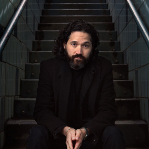 GOOD PEOPLE WITH MARK SAKAMOTO Image of a man wearing a black shirt and blazer with a large beard. He is sitting on steps.
