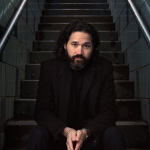 Image of a man wearing a black shirt and blazer with a large beard. He is sitting on steps.