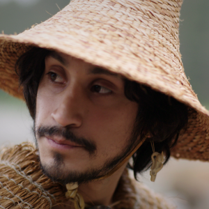 SG̲AAWAAY Ḵ’UUNA (EDGE OF THE KNIFE) Close up photo of a man wearing a straw hat