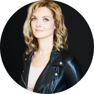 Blonde woman with blue eyes wears a cream coloured top and black leather jacket standing in front of a black background