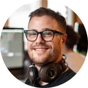 Man with short brown hair and thick rimmed glasses smiles at the camera. He has big headphones around his neck.